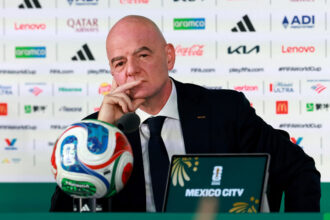 Bald man in a suit at a press conference, with sponsor logos in the backdrop and a microphone in front, Mexico City visible on a sign.