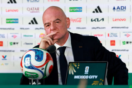 Bald man in a suit at a press conference, with sponsor logos in the backdrop and a microphone in front, Mexico City visible on a sign.