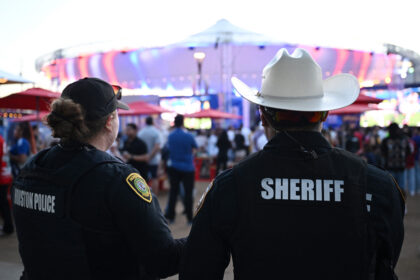 Two sheriff officers stand with their backs to the camera at an outdoor event, overlooking a crowd and a brightly lit stage stage in the background.