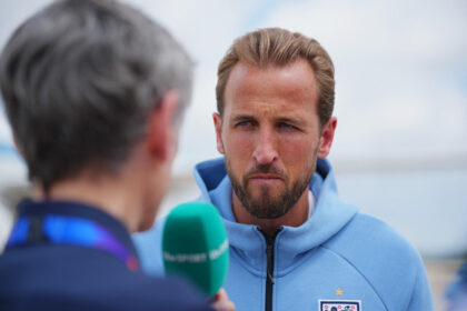 Man in a light blue jacket being interviewed outdoors, English football badge visible on chest, blurred interviewer with a green microphone in foreground