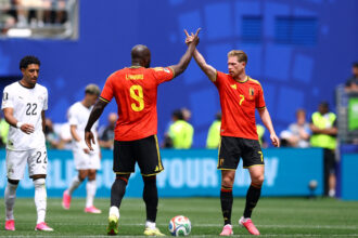 Belgian teammates in red celebrate with a high-five on the field, ball at their feet, blue stadium backdrop behind them