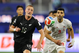 Two male soccer players race for the ball during a match, one in a black kit and the other in white, with the ball between them.