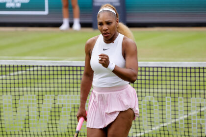 Tennis player in a white tank top and pink skirt walks toward the net, gripping a pink racket outside on a tennis court.