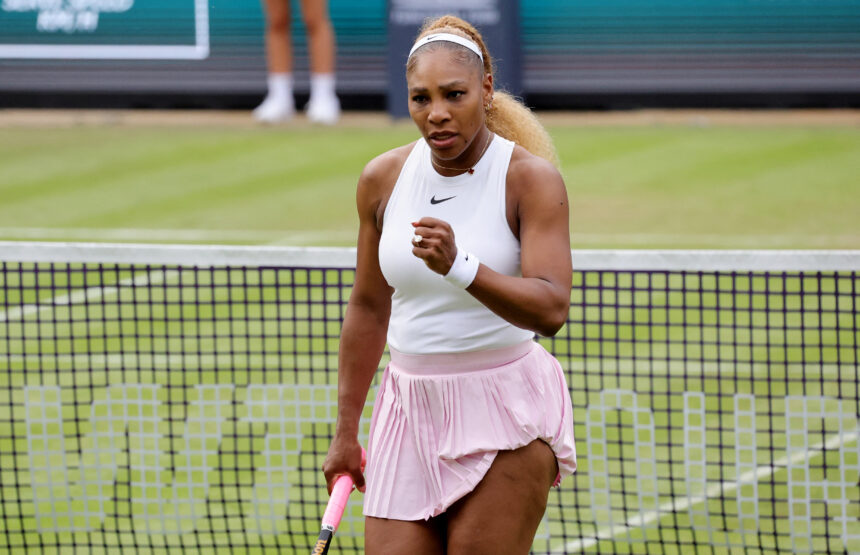 Tennis player in a white tank top and pink skirt walks toward the net, gripping a pink racket outside on a tennis court.