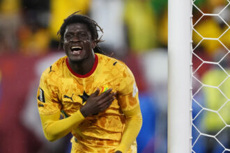 Soccer goalkeeper in a yellow patterned jersey celebrating near the goal net, hand on chest.