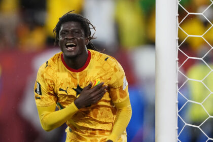 Soccer goalkeeper in a yellow patterned jersey celebrating near the goal net, hand on chest.