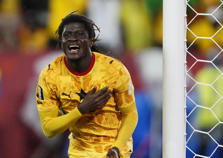 Soccer goalkeeper in a yellow patterned jersey celebrating near the goal net, hand on chest.
