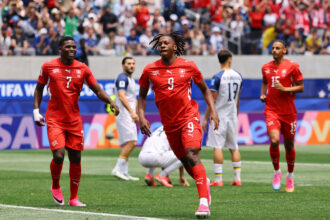 Soccer players in red uniforms sprint toward the camera on a green pitch as a crowded stadium watches from the stands.