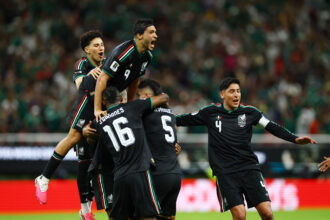 Mexican national team players celebrate a goal, jumping and embracing on the field as cheering fans blurred in the background.