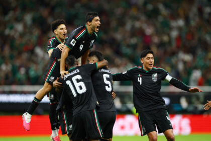 Mexican national team players celebrate a goal, jumping and embracing on the field as cheering fans blurred in the background.