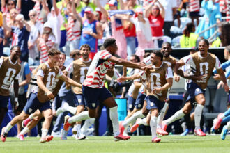 Group of USA soccer players in brown bibs running across a green field as fans cheer in the stands.