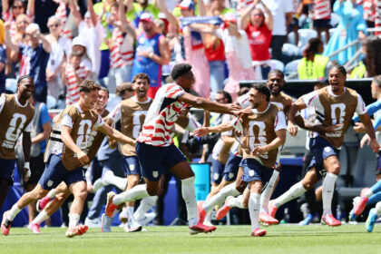 Group of USA soccer players in brown bibs running across a green field as fans cheer in the stands.