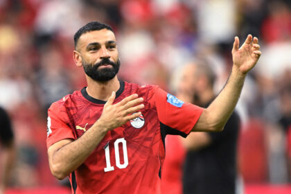 Egyptian footballer in a red jersey with number 10, raising his hand in celebration on the field.