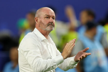 Man in a white shirt clapping on the sidelines, blurred blue stadium seats in the background.