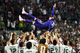 Soccer team in white jerseys lifts a teammate in blue into the air as a cheering crowd looks on in a stadium.