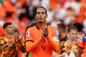 Soccer player in an orange Netherlands jersey with number 4 clapping on the field, stands filled with fans in orange behind him.
