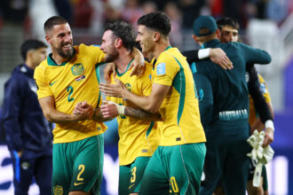 Australian soccer players in yellow and green celebrate with arms around each other on the field.