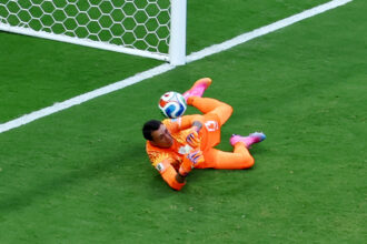 Soccer goalkeeper in orange saves a ball near the goal on a green field.