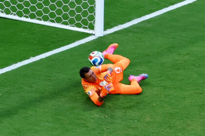 Soccer goalkeeper in orange saves a ball near the goal on a green field.