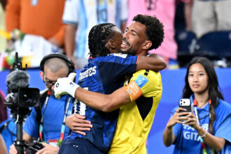 Two soccer players in blue and yellow hug in celebration as a photographer records the moment with a video camera nearby.
