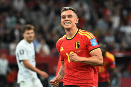 Belgian footballer in red jersey celebrates on the pitch during a match, fists clenched and smiling, crowd blurred in background