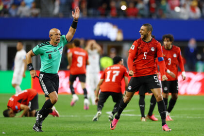 Soccer players in red celebrate a goal on the green pitch as a teal referee raises his arm in celebration gesture behind them.
