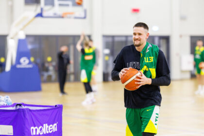 Basketball player in green-yellow uniform holding a ball in a gym, smiling, with blurred teammates in the background and a purple barrier in the foreground.