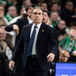 Man in a dark suit and blue tie stands in a crowded sports arena, gesturing with his right arm during a game banter moment.”,
