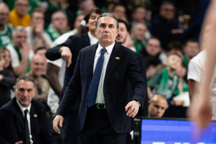Man in a dark suit and blue tie stands in a crowded sports arena, gesturing with his right arm during a game banter moment.”,