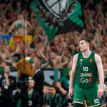 Basketball player in a green Zalgiris jersey number 10 walking on court with a cheering crowd in the stands behind him.