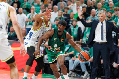 Two basketball players battle for control near the baseline as a coach in a suit raises his arm in the background and fans watch from the stands in green.