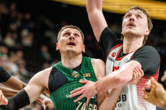 Two basketball players in green and white jerseys clash for the ball, arms locked amid a crowded court.