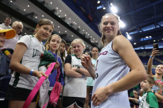 Female athlete in a white jersey signs autographs for a crowd of young fans at a sports event in an arena.