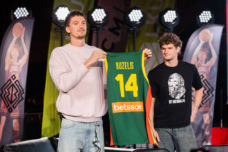 Two men on a stage hold up a green and yellow basketball jersey that reads BUZELIS 14; stage lights and banners in the background.