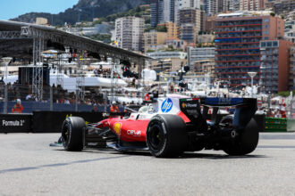 Red Formula 1 race car on a track with city buildings and scaffolding in the background, sponsor logos visible on the car