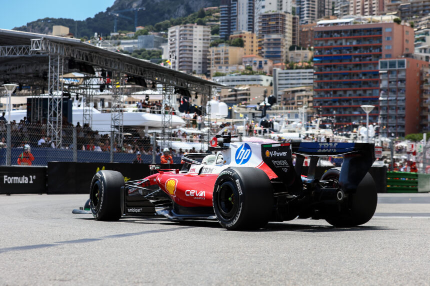 Red Formula 1 race car on a track with city buildings and scaffolding in the background, sponsor logos visible on the car