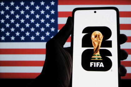 Hand holding a smartphone displaying the FIFA World Cup trophy icon in front of an American flag background.