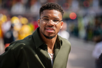 Close-up portrait of a man with short curly hair and clear-framed glasses, wearing a green button-up shirt, outdoors and looking at the camera.