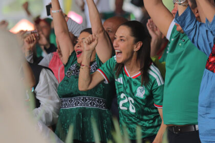 Crowd of cheering sports fans with raised arms; woman in a green Mexico jersey smiles in the foreground.