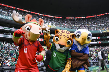 Four colorful sports mascots pose at the edge of a stadium field as a large, cheering crowd fills the stands behind them, wearing red, green, and blue jerseys.