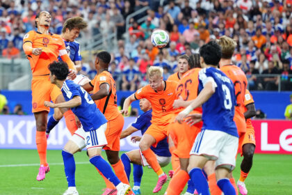 Soccer players in orange and blue jerseys jump to head the ball amid a crowded stadium.