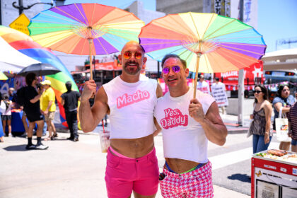 Two men posing at a Pride parade under rainbow umbrellas; one in pink shorts with a 'Macho' shirt, the other in a white 'Yes Daddy' shirt and pink sunglasses.