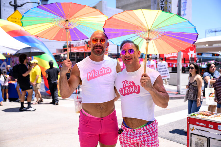 Two men posing at a Pride parade under rainbow umbrellas; one in pink shorts with a 'Macho' shirt, the other in a white 'Yes Daddy' shirt and pink sunglasses.