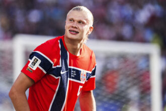 Male athlete in a red, white, and blue Norwegian jersey, looking to the left on a sports field.