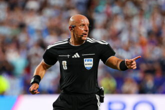 Bald soccer referee in black kit with FIFA badge and headset on a crowded field, gesturing as play continues.