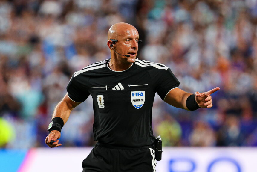 Bald soccer referee in black kit with FIFA badge and headset on a crowded field, gesturing as play continues.