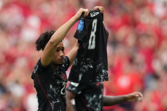 Two players lift a black jersey overhead in celebration on a crowded stadium filled with red-clad fans.