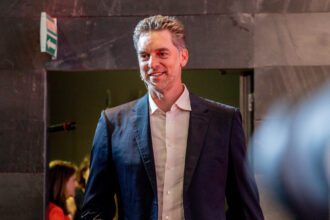 Smiling man in a navy blazer and white shirt stands in a dim venue with an exit sign in the background.