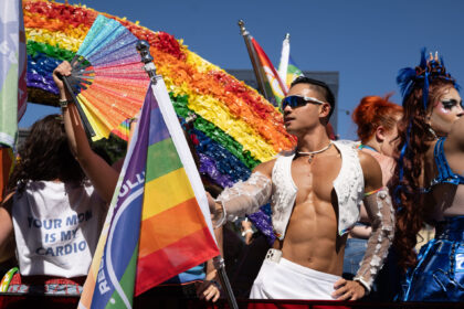 People at a Pride parade waving rainbow flags under a clear blue sky, wearing colorful outfits and makeup.