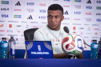 Male footballer at a press conference, wearing a light shirt and chain, in front of a sponsor wall with logos and a microphone.